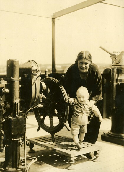 En vuxen och ett litet barn står vid ett stort skeppsratt på ett fartygsdäck, sepiafärgad bild.