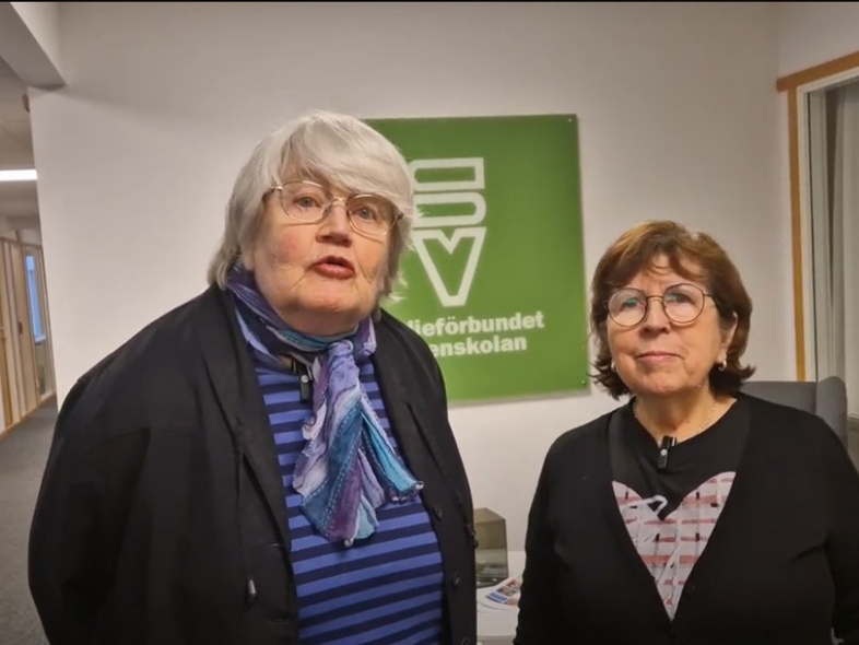 Två äldre kvinnor står inomhus framför en grön skylt med text, båda tittar mot kameran.