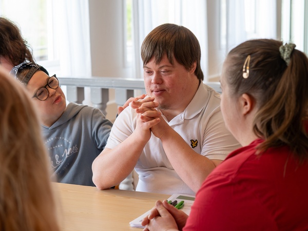 En grupp unga vuxna med Downs syndrom sitter runt ett bord och samtalar i ett ljust rum.