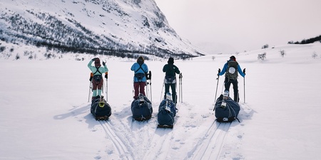 Fyra personer med pulkor står på skidor i ett snötäckt fjällandskap med berg i bakgrunden.