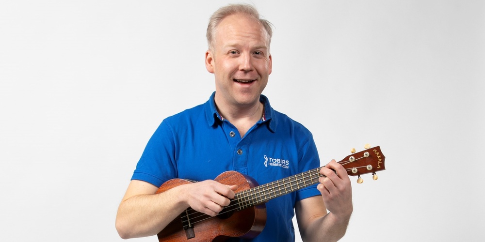 Man i blå pikétröja spelar ukulele och står mot en vit bakgrund.