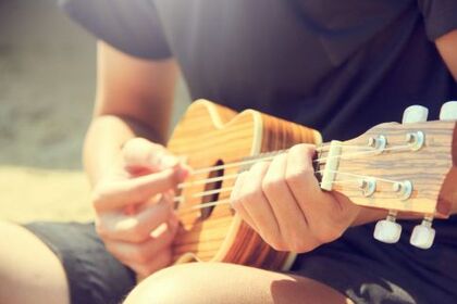 En person spelar ukulele utomhus i solljus, fokus på händer och instrument.
