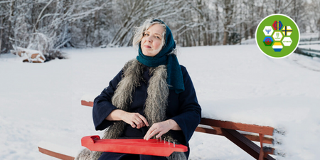 Kvinna i vinterkläder sitter på bänk i snölandskap och spelar ett rött stränginstrument.