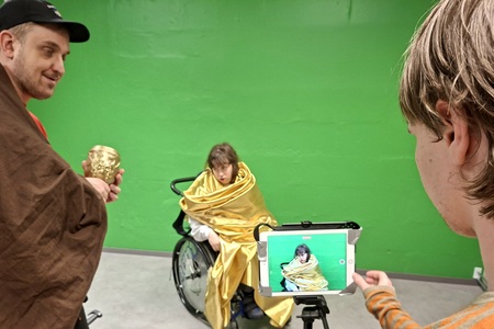Tre personer spelar in en film framför en grön vägg, en sitter i rullstol med gul mantel, en filmar med surfplatta.