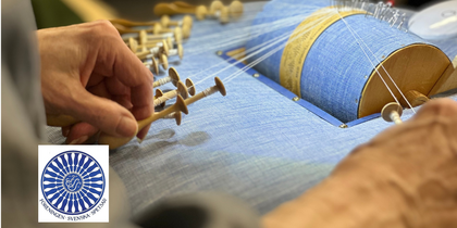 Närbild av händer som arbetar med knyppling på en blå knyppeldyna, med flera trådar och pinnar.