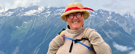 Leende kvinna med hatt och ryggsäck står på en grön sluttning med berg i bakgrunden.