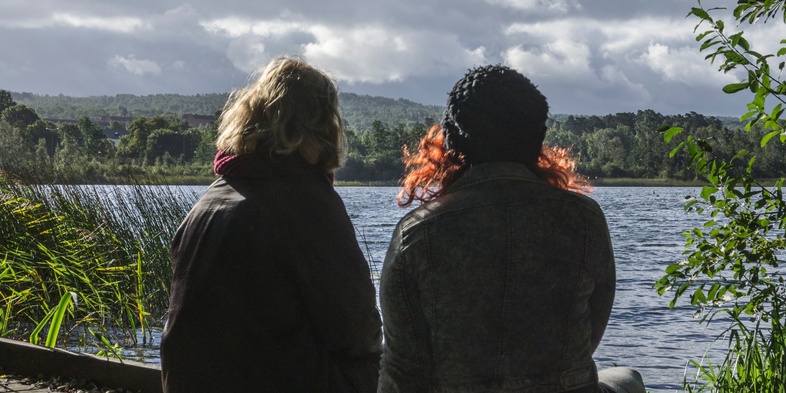 Två personer sitter på en brygga vid en sjö och tittar ut över vattnet under en molnig himmel.