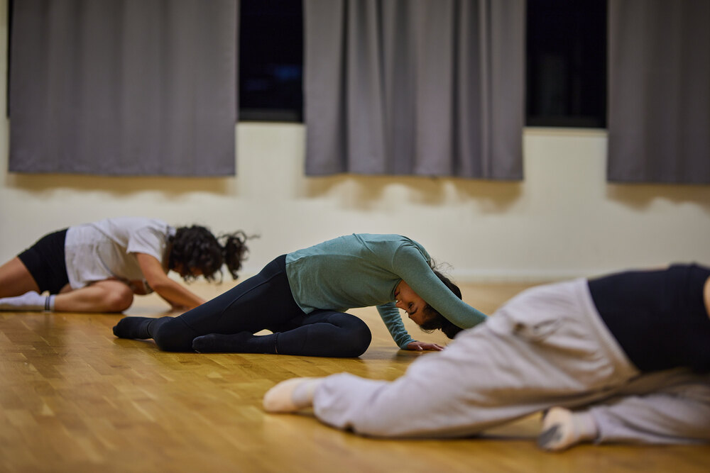Tre personer gör stretchövningar på golvet i en dansstudio med trägolv och grå gardiner.