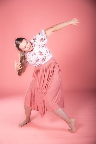 Person i blommig topp och rosa kjol poserar barfota mot en rosa bakgrund.