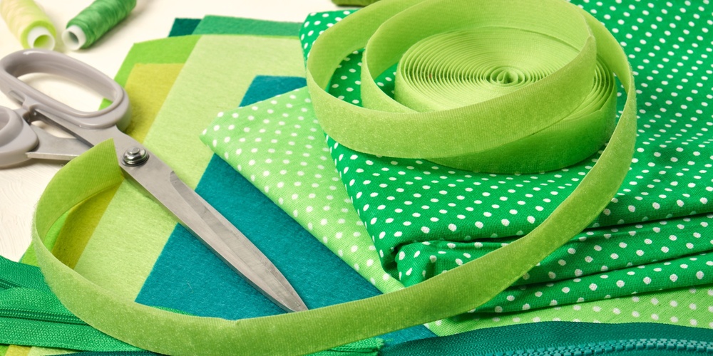 Sytillbehör i gröna nyanser, inklusive tyg, sax, dragkedja, tråd och band, ligger utspridda på ett bord.