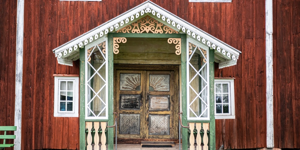 Entré till ett rött trähus med snickarglädje, gröna detaljer och dubbeldörrar i trä.