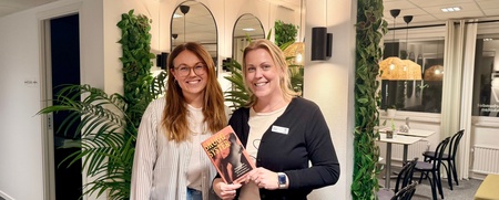 Två kvinnor står och ler i ett modernt kontorsrum, en håller en bok med titeln 'Succé Syster'.