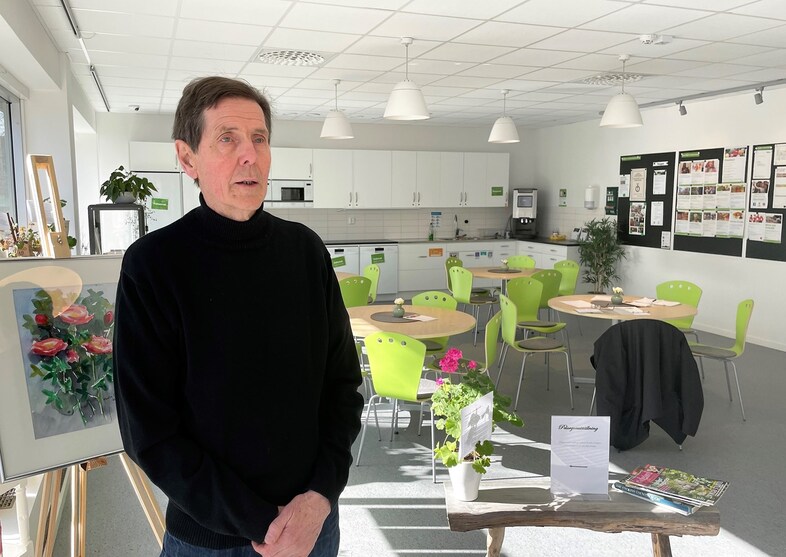 En man i svart tröja står i ett ljust, modernt rum med gröna stolar, runda bord och växter.