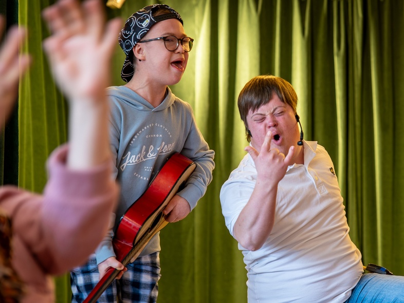 Två unga personer, en med gitarr, sjunger och gör rockposer framför gröna gardiner.