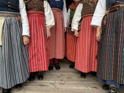 Flera personer bär traditionella folkdräkter med randiga förkläden i olika färger, stående på ett trägolv.