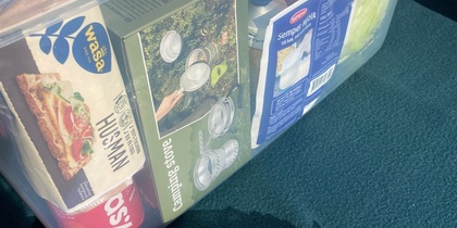 En genomskinlig plastlåda fylld med matvaror och hushållsartiklar står på ett bord utomhus.