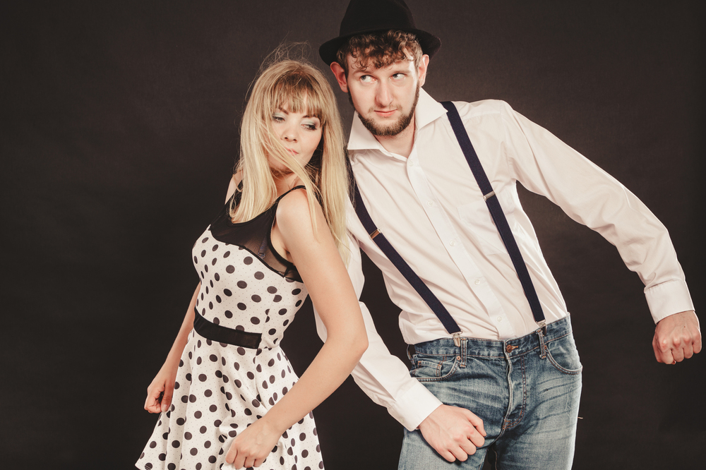 En kvinna i prickig klänning och en man i hatt och hängslen dansar tillsammans mot mörk bakgrund.