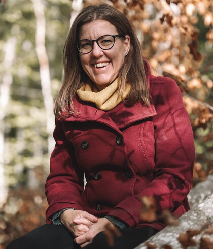 Kvinna med glasögon och röd kappa sitter utomhus bland höstlöv och ler mot kameran.