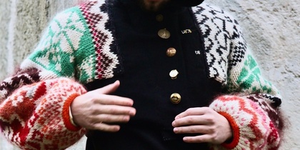 Person i svart huvtröja med färgglada stickade ärmar och guldknappar står mot en betongvägg.