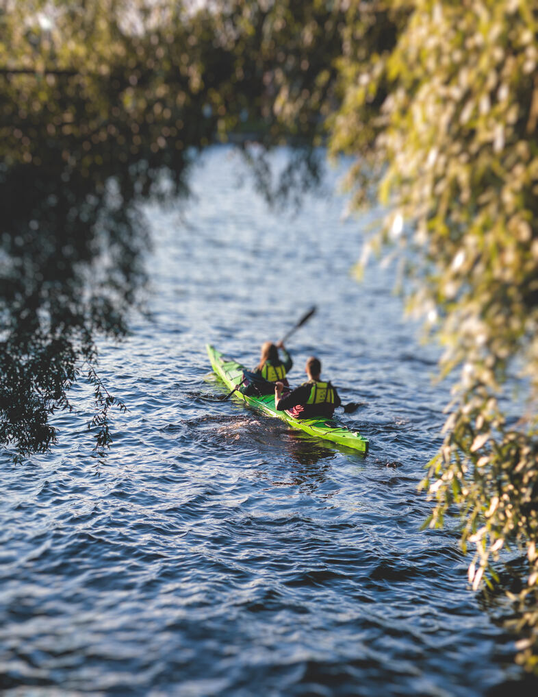 Två personer paddlar en grön kajak på en flod omgiven av trädgrenar.