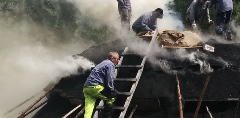 Flera personer arbetar på en rykande kolmila utomhus, omgivna av skog och tjock rök.