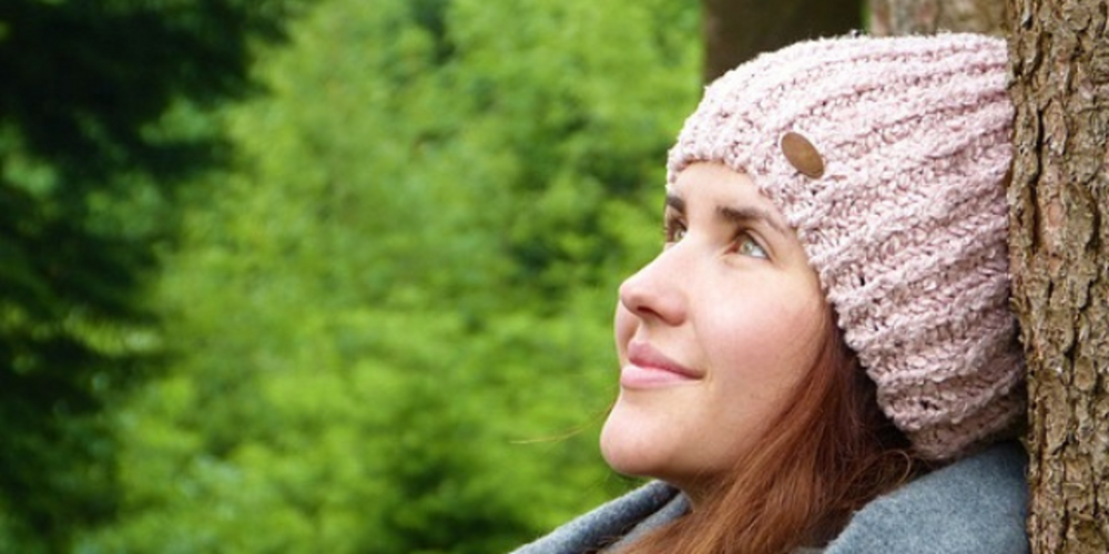 Person med stickad mössa och halsduk lutar mot ett träd i en grön skogsmiljö.
