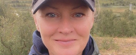 Kvinna i grön jacka och svart keps tar en selfie utomhus i naturmiljö.