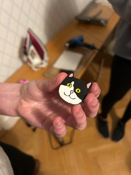 En hand håller en liten figur av en svartvit katt med gula ögon, i ett rum med trägolv och strykjärn i bakgrunden.