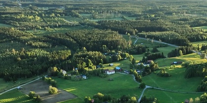 Flygfoto över ett grönt landskap med åkrar, skog och utspridda hus i kvällsljus.