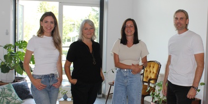 Fyra personer står i ett ljust vardagsrum med växter och stora fönster.