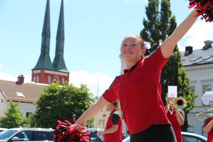 En ung cheerleader i röd tröja ler och håller pom-poms utomhus, med en kyrka i bakgrunden.