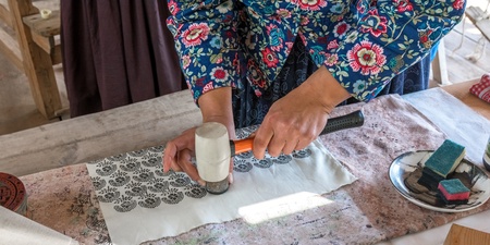 En person trycker mönster på tyg med hjälp av en stämpel och hammare vid ett bord.
