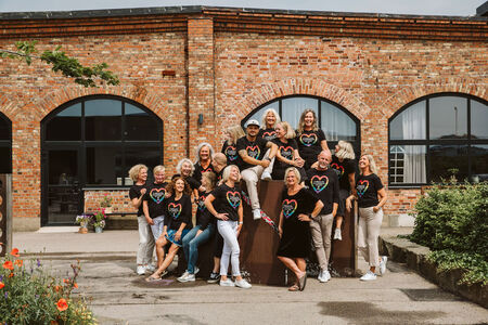 En grupp glada människor i svarta t-shirts med hjärtmotiv poserar utomhus framför en tegelbyggnad.
