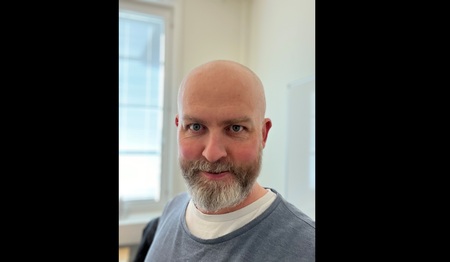 En skallig man med skägg står inomhus framför ett fönster och tittar in i kameran.