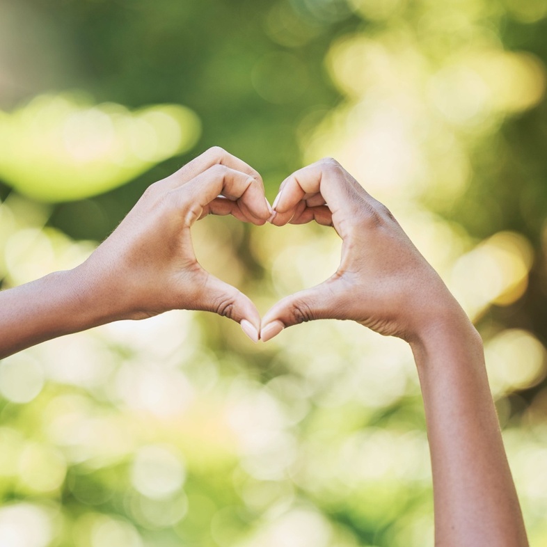Två händer formar ett hjärta mot en suddig, grön bakgrund utomhus.