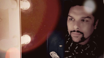 Man med skägg och scarf sitter vid en spegel med lampor, mjukt ljus och varma färger.