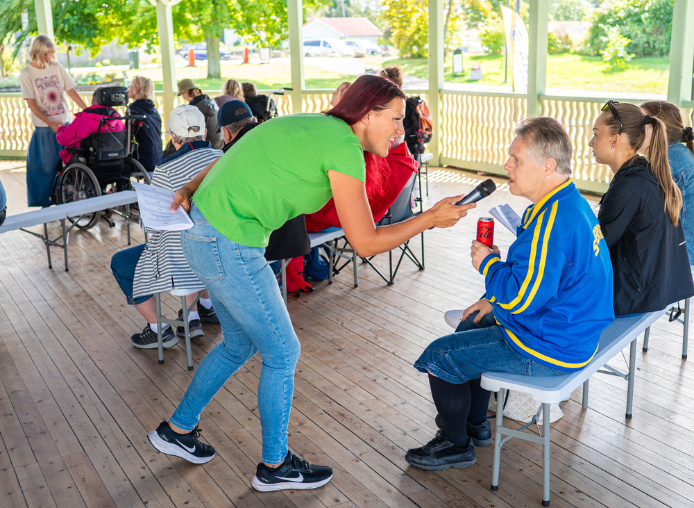 En kvinna intervjuar en man med mikrofon i en paviljong med flera sittande personer.