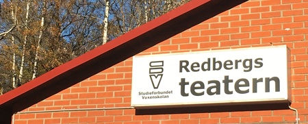 Röd tegelbyggnad med skylt där det står 'Redbergs teatern', träd och blå himmel i bakgrunden.