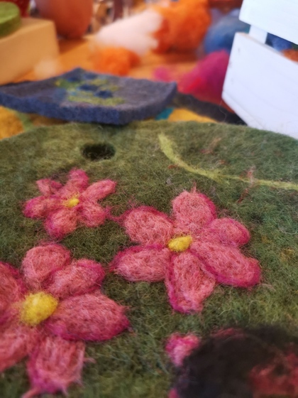 Närbild på tovade ullblommor i rosa och grönt, med suddig bakgrund av hantverksmaterial.