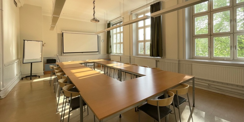 Ljust klassrum med stora fönster, U-formade bord och stolar, whiteboard och blädderblock.