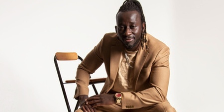 Person i beige kostym sitter på en stol mot vit bakgrund, ansiktet är suddat.