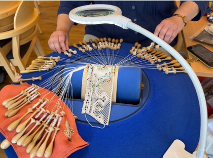 En person tillverkar spets med hjälp av knyppelpinnar, tråd och en förstoringslampa på ett blått bord.