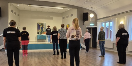 En grupp personer står i ett dansrum och följer en instruktör på en liten scen längst fram.