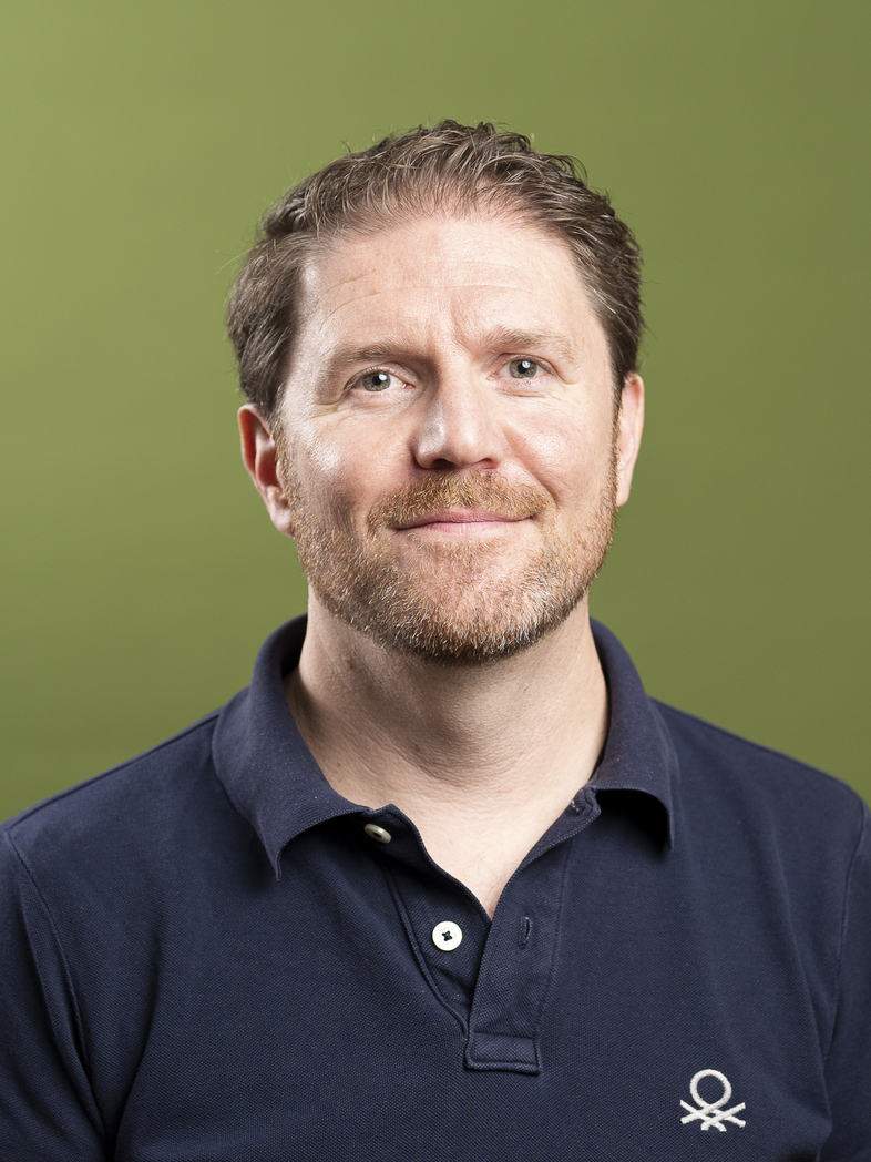 Man i mörkblå pikétröja står mot en grön enfärgad bakgrund.