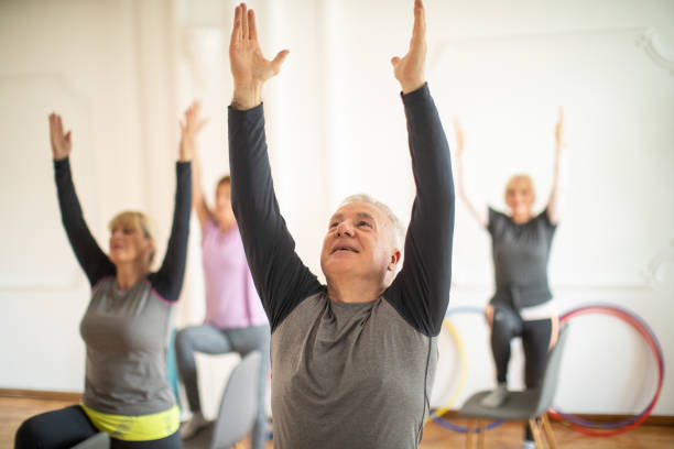 Äldre personer gör sittande yogaövningar med armarna uppsträckta i ett ljust rum.