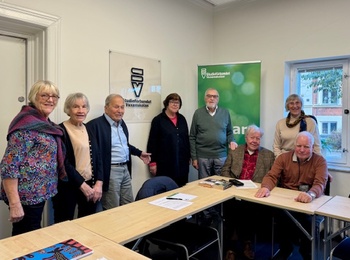 En grupp äldre personer står och sitter runt ett konferensbord i ett mötesrum.