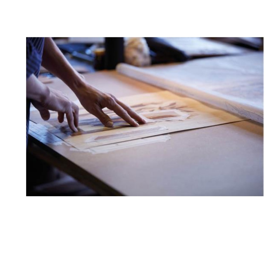 Två händer arbetar med ritningar eller mönster på ett bord i ett ljust rum.