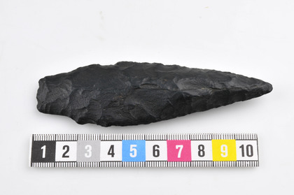 Svart stenålderspilspets av flinta, liggande bredvid en färgad linjal på vit bakgrund.