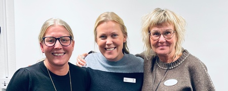 Tre personer står tillsammans i ett ljust rum, klädda i stickade tröjor och med namnskyltar.