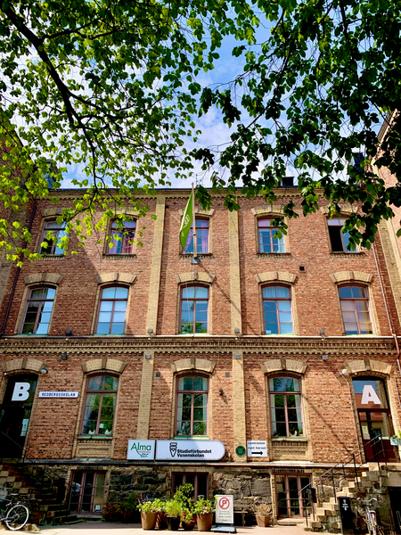 Tegelfasad på en äldre byggnad med skyltar, gröna trädgrenar i förgrunden och blå himmel ovanför.